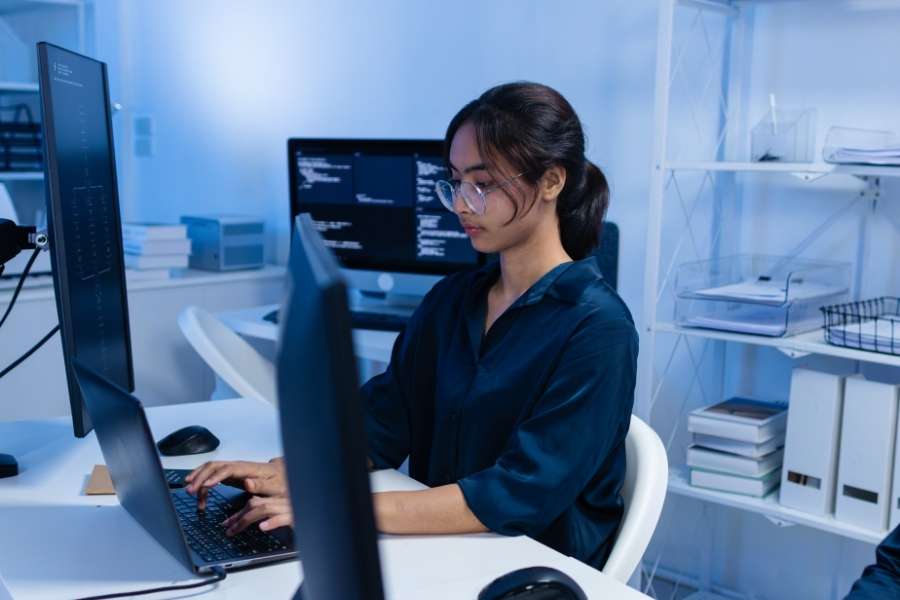Une informaticienne travaille sur son ordinateur portable dans un bureau équipé de plusieurs écrans.