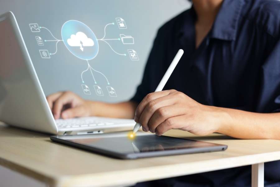 Une personne utilise un stylet sur une tablette tactile à côté d'un ordinateur portable, illustrant la gestion des bases de données et le stockage cloud.